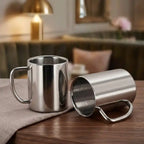 Two stainless steel mugs on a wooden table with a blurred indoor background